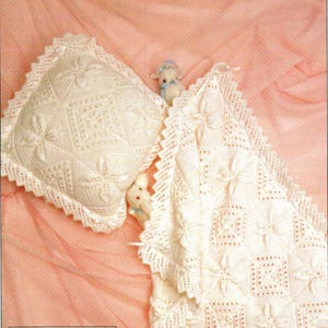 May include: Cream-colored, crocheted baby blanket and pillow set. The blanket and pillow feature a floral and geometric pattern with a scalloped edge. Two small stuffed animals are nestled in the blanket. The set is on a pink background.