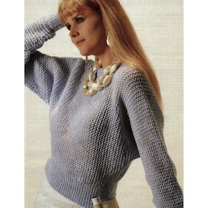 May include: A pale blue knitted sweater with a textured design. The sweater features long sleeves and a ribbed hem. The image shows a woman wearing the sweater and a large, off-white necklace.