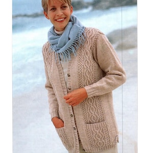 May include: A beige cable-knit cardigan with a button-up front and two front pockets. The cardigan features a textured cable design on the front panels and sleeves. A light blue scarf is worn around the neck.