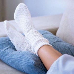 Puede incluir: Primer plano de calcetines blancos de punto con puños acanalados. Los calcetines están en las piernas de una persona, descansando sobre una manta azul de punto. Los calcetines parecen suaves y cómodos, sugiriendo un ambiente relajado.