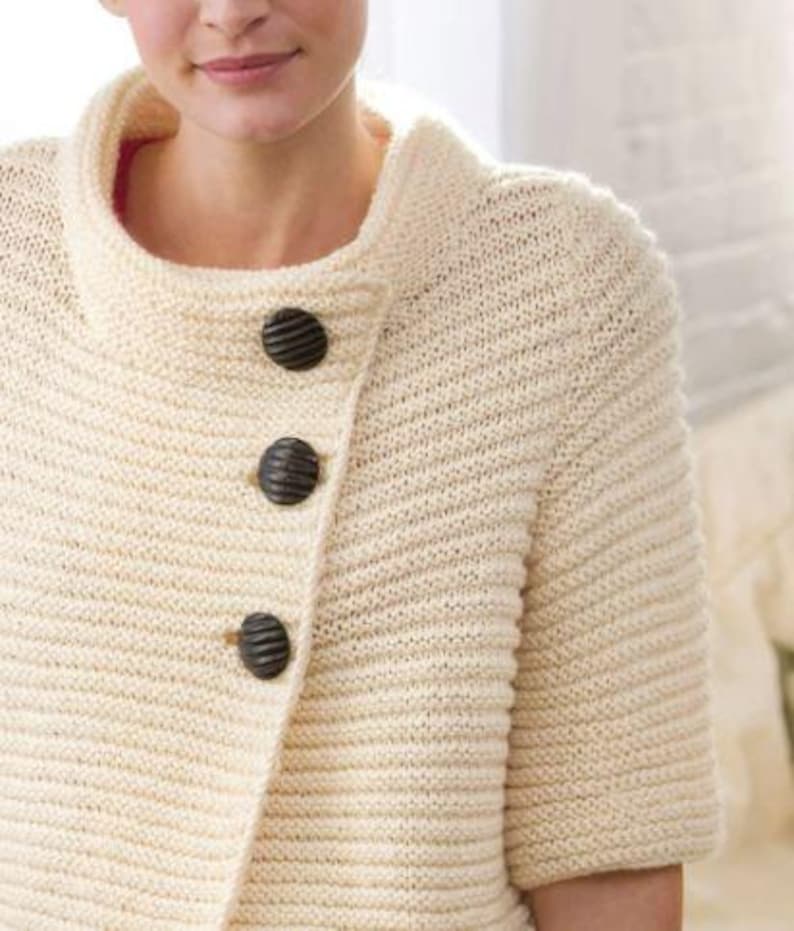 Pu&ograve; includere: Un cardigan di maglia color crema con chiusura leggermente asimmetrica e tre bottoni marroni scuri. Il cardigan ha un collo alto e maniche corte.