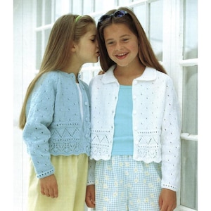 Puede incluir: Dos chicas jóvenes con cárdigans de punto, uno azul claro y otro blanco. Ambos tienen cierres de botón y diseños calados decorativos. Las chicas están de pie frente a una ventana blanca.