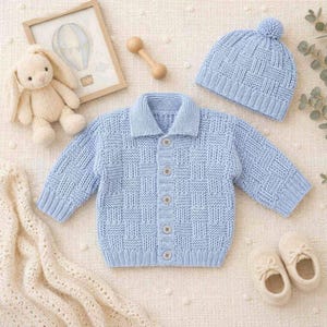 May include: A baby's light blue knitted outfit set, featuring a button-down cardigan, a beanie with a pom-pom, and matching booties. The set is displayed with a stuffed bunny, a framed picture, and a wooden rattle.