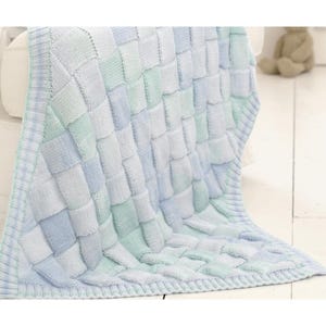 Puede incluir: Una manta de punto en tonos azules, verdes y blancos, con un patrón de tejido de cesta. La manta está drapeada sobre una superficie blanca, con un osito de peluche visible en el fondo.