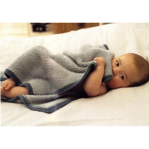 Puede incluir: Un bebé envuelto en una manta gris tejida con un borde azul, acostado sobre una sábana blanca. La manta es texturizada y cubre la mayor parte del cuerpo del bebé, con la cara y un brazo visibles. El bebé mira a la cámara.