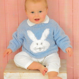 May include: A baby in a light blue knitted sweater featuring a white bunny face. The sweater has a white collar and long sleeves. The baby is also wearing white trousers. The background is a pink wooden surface.