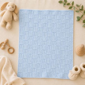 May include: A light blue knitted blanket featuring a textured basketweave pattern. The rectangular blanket is surrounded by a beige plush rabbit toy, a wooden rattle, a wooden stacking toy, a pair of beige knit booties, and a piece of cream fabric.