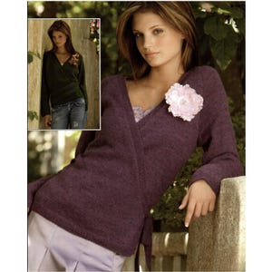 Può includere: Un maglione avvolgente viola scuro con maniche lunghe e scollo a V. Un grande fiore rosa chiaro è appuntato sul maglione. Il maglione è legato in vita. Un'immagine più piccola mostra un maglione verde simile.