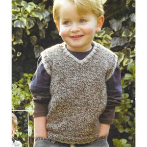 Op de afbeelding: Een kind draagt een bruin en wit gespikkelde gebreide vest met een V-hals. Het vest is gelaagd over een blauw shirt met lange mouwen. Het kind lacht voor een groene, bladerrijke achtergrond.