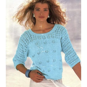 May include: A light blue, open-knit sweater with three-quarter sleeves. The sweater features a round neckline and a delicate, patterned design around the neckline and sleeves. A blue bracelet is visible on the left wrist.