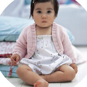 May include: A baby wearing a light pink knitted cardigan and a white romper with a floral and bunny print. The baby is sitting on a light-colored surface.