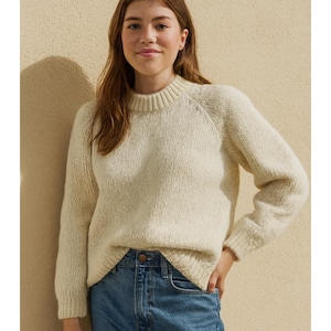 Könnte beinhalten: Cremefarbener Pullover mit geripptem Stehkragen und Raglanärmeln. Der Pullover ist aus einem strukturierten Strickstoff gefertigt und wird mit blauen Jeans kombiniert. Der Gesamtstil ist lässig und bequem.