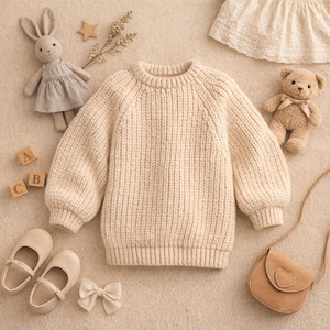 May include: Cream-colored knit sweater with a round neck and long sleeves. The sweater is surrounded by children's accessories, including a stuffed bunny, a teddy bear, a small purse, shoes, and wooden blocks.