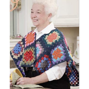 May include: A colorful, handmade crochet shawl featuring a patchwork design. The shawl is composed of various square motifs in shades of blue, red, orange, and green. The garment is worn over a white collared shirt and black pants.