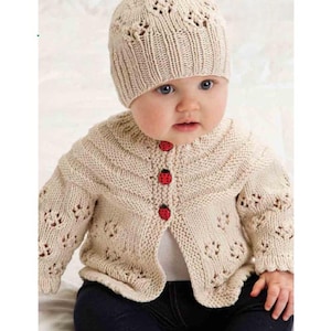 May include: A beige knitted baby cardigan and matching hat. The cardigan has red ladybug buttons and decorative openwork. The baby is wearing dark blue pants. The hat has a scalloped edge and openwork design.