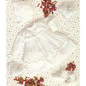 May include: A white knitted baby outfit set, comprising a jacket, bonnet, and booties. The jacket features a scalloped hem and button closure. The set is arranged on floral fabric with dried red flowers and lace details.