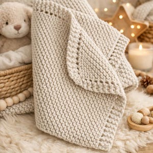 May include: A folded, cream-colored knitted blanket with a decorative edge. The blanket is made of thick yarn and has a textured appearance. A teddy bear and a basket are visible in the background, suggesting a nursery or cozy setting.
