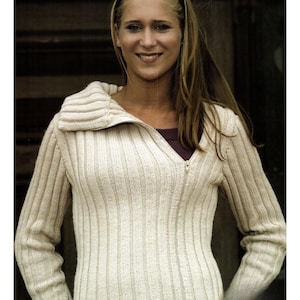 Puede incluir: Un suéter de punto acanalado de color crema con un cuello alto plegable y una cremallera de largo completo. El suéter tiene mangas largas y un patrón acanalado vertical. La modelo lleva una camisa morada oscura debajo.