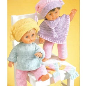 Peut inclure: Deux poupées vêtues de tenues tricotées. L'une porte un chapeau jaune, un pull bleu clair et un pantalon rose. L'autre porte un chapeau rose et violet, un poncho violet et un pantalon rose. Une écharpe rayée est posée sur la chaise.