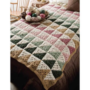 Puede incluir: Una manta de ganchillo hecha a mano con un patrón geométrico de triángulos en tonos blanco, rosa, verde y beige. La manta está sobre una cama con una colcha marrón. El borde es de color beige liso.