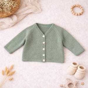 Puede incluir: Un cárdigan de bebé tejido de color verde salvia con cuello en V y mangas largas. El cárdigan tiene cinco botones blancos. También hay unos patucos de bebé color crema y un juguete de madera en la imagen.