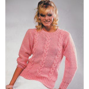Può includere: Un maglione rosa chiaro lavorato a maglia con girocollo e maniche lunghe. Il maglione presenta un motivo a trecce sulla parte anteriore. L'orlo e i polsini hanno una trama a coste. Il maglione è di un colore pastello tenue.