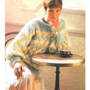 Może przedstawiać: Sweter z dzianiny z rombowym wzorem w pastelowych kolorach żółtym, niebieskim i białym. Sweter ma okrągły dekolt i długie, pękate rękawy. Mały, okrągły stolik ze złotą obwódką trzyma filiżankę kawy.
