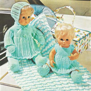 May include: Two vintage dolls in hand-knitted outfits. One doll is wearing a hooded jacket and trousers, the other a dress. Both are in a light blue colour. They sit on a matching blanket with a blue and white pattern. A baby carriage and a patterned bag are in the background.