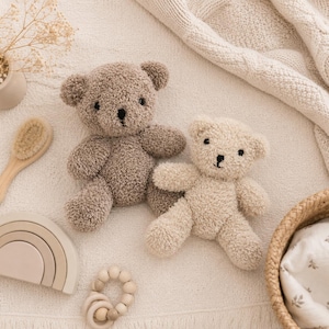 Può includere: Due orsacchiotti di peluche con una superficie soffice e testurizzata. Un orsetto è marrone chiaro e l'altro è color crema. Entrambi presentano semplici occhi e nasi neri ricamati. Sono disposti insieme su una superficie in tessuto dai toni neutri.