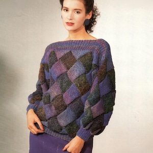 May include: A knitted sweater with a geometric pattern in shades of purple, blue, green, and brown. The sweater has a wide neckline and long sleeves. The model is wearing a purple skirt.