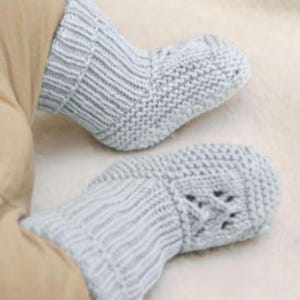 May include: Close-up of a pair of light gray knitted baby booties and mittens. The booties have a ribbed cuff and a textured knit pattern. The mittens feature a decorative openwork design. The items are made of soft yarn.