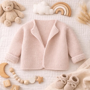 May include: A light pink knitted baby jacket with a shawl collar and folded cuffs. The jacket is surrounded by baby accessories, including a stuffed bunny, wooden rainbow, cloud, and baby shoes. The jacket is made of a soft, textured knit.