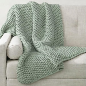 May include: A sage green knitted throw blanket draped over a light-coloured sofa. The blanket has a textured, chunky knit pattern. A white bolster pillow and a sofa arm are visible. The blanket appears soft and cosy, suitable for home decor.