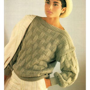 May include: A sage green knitted sweater with a textured, geometric pattern. The sweater has a wide neckline and long sleeves. The model is wearing a light-coloured hat and a light-coloured long-sleeved shirt underneath the sweater.