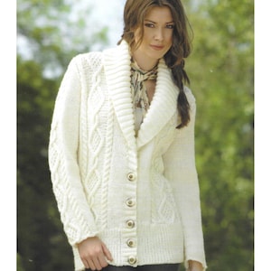 May include: A cream-coloured cable-knit cardigan with a shawl collar and gold-toned buttons. The sweater features intricate cable patterns and a ribbed collar. A woman is wearing the cardigan. The background is a soft green.