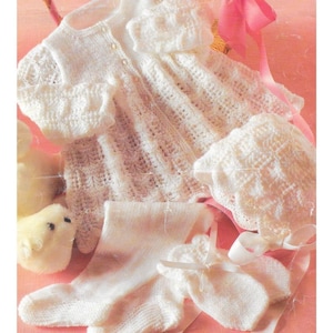 Könnte beinhalten: Ein weißes, gestricktes Baby-Outfit-Set, bestehend aus einer Strickjacke, einer Mütze, Socken und Fäustlingen. Das Set hat ein zartes Lochmuster und ist auf einer rosa Oberfläche angeordnet. Die Strickjacke hat kleine Knöpfe und wird mit einem rosa Band gebunden.