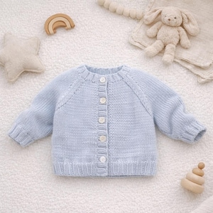May include: A light blue knitted baby cardigan with long sleeves and a round neckline. The cardigan has a button-up front with white buttons. A star-shaped pillow, a wooden rainbow, and a stuffed bunny are in the background.
