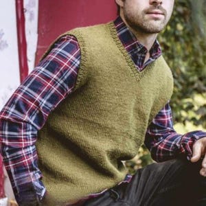 May include: A knitted olive green V-neck vest layered over a plaid button-down shirt with red, white, and blue accents. The vest has a textured knit pattern. The shirt has long sleeves.