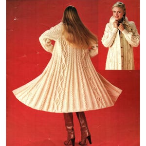 May include: Cream-colored, long knitted coat with a cable knit design. The coat has a flared skirt and long sleeves. The image also shows a close-up of the coat's collar and button closure. The model is wearing brown boots.
