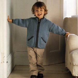 Könnte beinhalten: Eine blaue Strickjacke mit Reißverschluss, dunkelgrauen Verzierungen und Taschen. Die Jacke wird mit einer khakifarbenen Hose getragen. Das Kind lächelt und steht in einem Türrahmen.