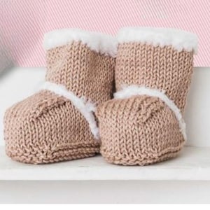 May include: A pair of knitted baby booties in a light brown color, trimmed with white faux fur around the top and ankle. The booties are made of a thick, textured yarn and sit on a white surface.