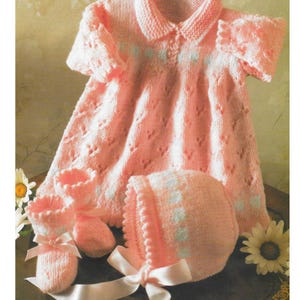 Pode incluir: Um conjunto de bebé tricotado em rosa claro, incluindo um vestido, um gorro e botinhas. O vestido tem gola e mangas curtas. O gorro e as botinhas têm detalhes brancos e laços de fita de cetim. Um conjunto delicado e charmoso.