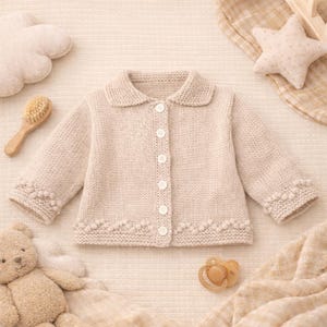 Puede incluir: Cárdigan de bebé tejido de color beige claro con cuello redondo y cierre de botones. Las mangas y el dobladillo presentan un punto decorativo de bolitas. El cárdigan está rodeado de artículos para bebés, como un oso de peluche.