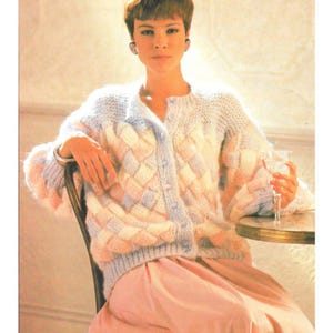 Könnte beinhalten: Ein pastellfarbener Strickcardigan mit einem Korbgeflechtmuster. Der Cardigan hat eine Knopfleiste vorne und lange Ärmel. Die Person im Bild trägt einen rosa Rock und sitzt auf einem Stuhl.