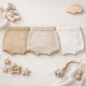 Può includere: Tre bloomers per neonati lavorati a maglia in beige, crema e bianco sono esposti su una superficie bianca. Sono presenti anche un coniglietto di peluche, stelle di legno, una nuvola e un ciuccio con una catena di perline di legno, che suggeriscono un tema per la cameretta.