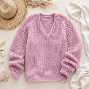 Può includere: Un maglione rosa chiaro lavorato a maglia con scollo a V e maniche lunghe. Il maglione ha una trama a coste e una vestibilità morbida. Le maniche sono raccolte ai polsini. Il maglione è esposto su una superficie neutra con un cappello di paglia e conchiglie.