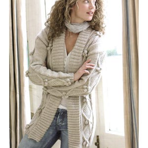 Könnte beinhalten: Beigefarbene Strickjacke mit langen Ärmeln und strukturiertem Strickmuster. Die Strickjacke hat einen gerippten Kragen und Bündchen, mit dekorativen Bommeln und Rautenmustern. Das Model trägt eine blaue Jeans und einen hellgrauen Schal.