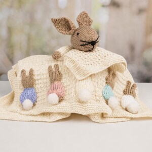 May include: A cream-colored knitted security blanket featuring a plush rabbit head at the top and five small knitted rabbit figures along the bottom edge. Each small rabbit has a white pom-pom tail and wears a different colored sweater in blue, pink, or teal.