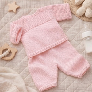 Può includere: Un completo neonato in maglia rosa chiaro, composto da un top a maniche lunghe e pantaloncini abbinati. Il completo è realizzato in filato morbido e viene esposto su una superficie trapuntata. Sono visibili anche una stella di legno e un biberon.