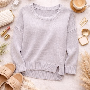 May include: A pale grey knitted jumper with a round neck and side slits. The jumper is surrounded by beauty products, a cup of coffee, and sandals. The jumper is made of a soft, comfortable material.